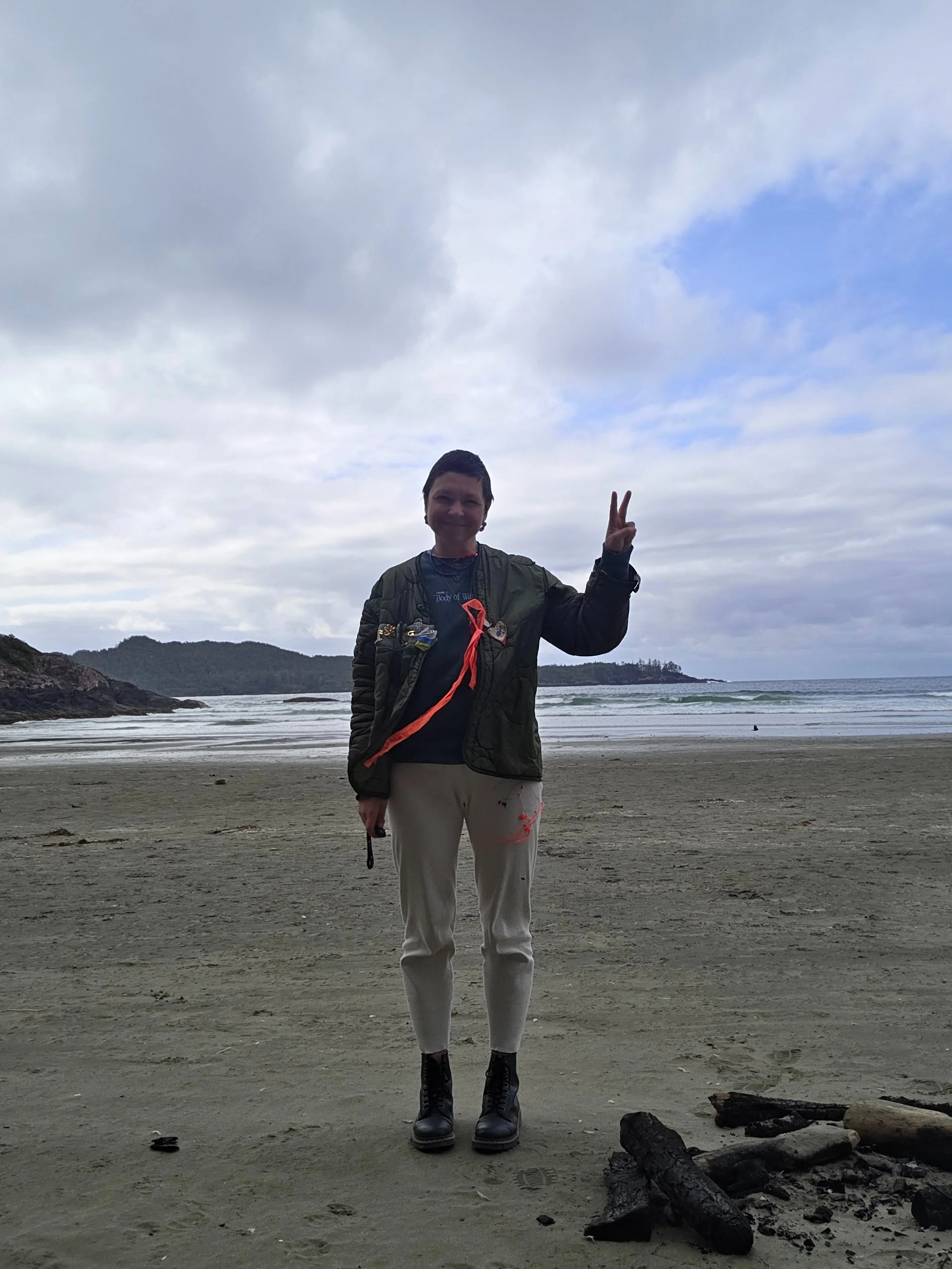 Varvara on a beach in a mended military jacket with pins, giving a peace sign